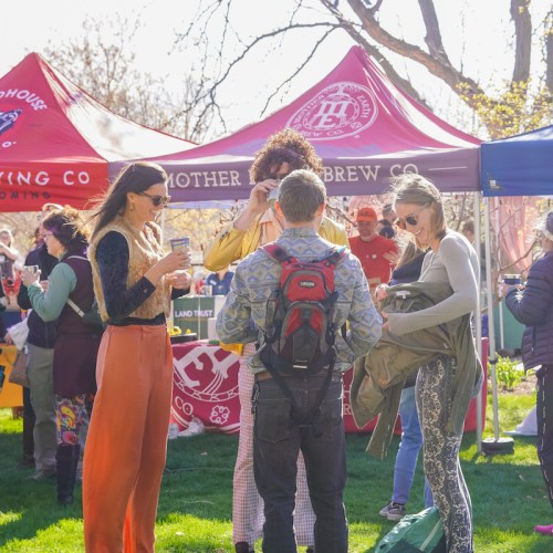 A group of people chatting at an outdoor event with colorful tents in the background, enjoying a sunny day.