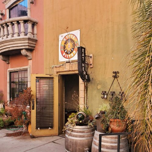 A colorful building entrance with a sign, surrounded by decorative barrels and plants.