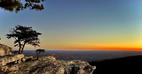 A serene sunset view from a rocky overlook, featuring silhouetted trees against a gradient sky.