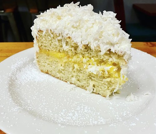 A slice of coconut cake topped with shredded coconut and dusted with powdered sugar on a white plate.
