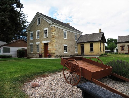 Historic stone house with a wooden cart in the foreground, surrounded by green grass and trees under a cloudy sky.