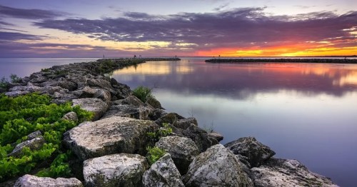 A rocky jetty extends into calm waters, reflecting a vibrant sunset with colorful clouds above.