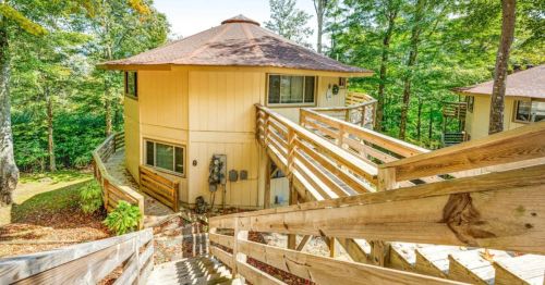 A wooden staircase leads down to a round, two-story cabin surrounded by trees and autumn foliage.