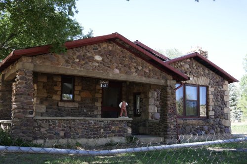 A stone house with a red roof and porch, surrounded by trees and a grassy area.