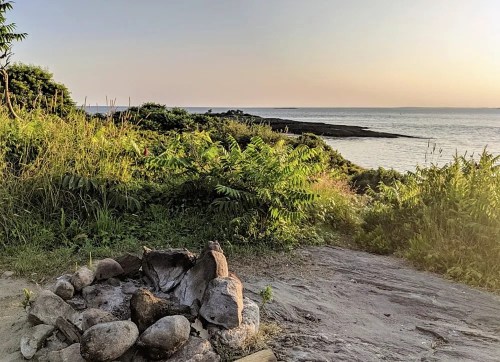 A serene coastal view at sunset, featuring a fire pit surrounded by lush greenery and calm waters in the background.