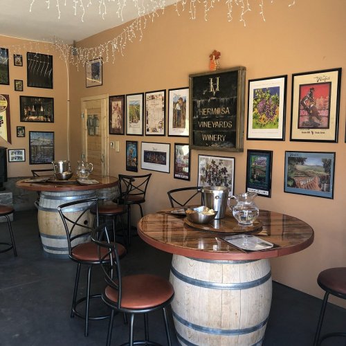 Cozy winery interior with wooden tables, barrel seating, and framed artwork on the walls. Warm, inviting atmosphere.