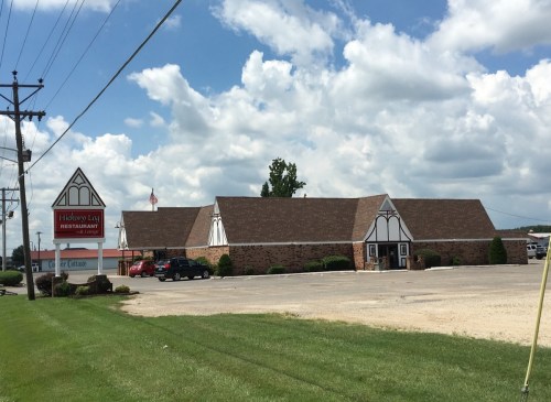 A large, rustic restaurant building with a sloped roof, surrounded by green grass and a cloudy sky.
