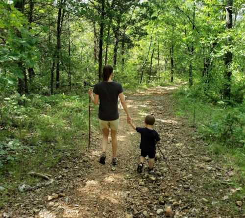 A woman and a child walk hand in hand along a forest trail, surrounded by lush greenery and trees.