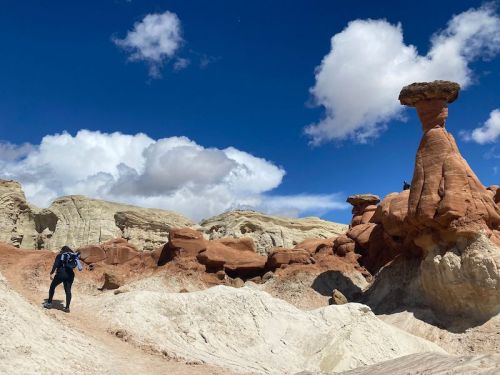 A hiker explores a colorful rock formation under a blue sky with fluffy clouds in a desert landscape.