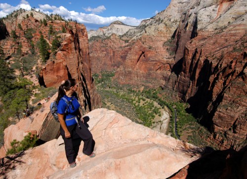 A hiker stands on a rocky ledge overlooking a deep canyon with red cliffs and a winding river below.