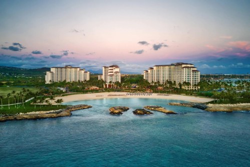 Aerial view of a beachfront resort with multiple buildings, palm trees, and calm waters under a colorful sky.