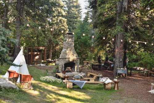 A cozy outdoor gathering space with a stone fireplace, seating, and string lights among tall trees.
