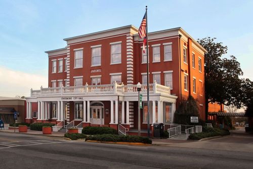 Historic red brick city hall building with white trim, American flag, and landscaped entrance in a small town.