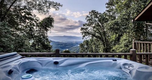 A hot tub on a deck overlooking a scenic mountain view with trees and clouds in the background.