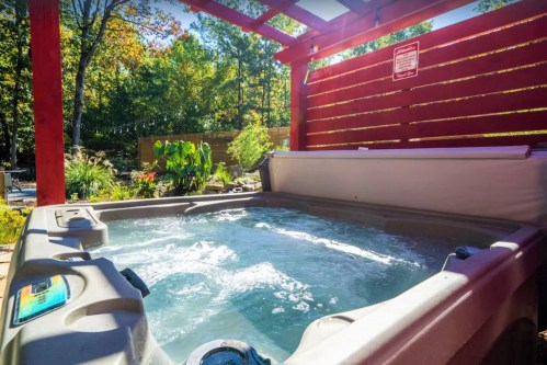 A hot tub surrounded by greenery and a wooden pergola, with sunlight filtering through the trees.