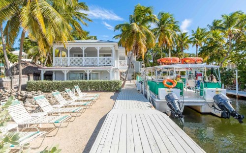 A tropical waterfront scene featuring a house, palm trees, lounge chairs, and boats docked by a wooden walkway.
