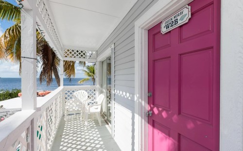 A bright pink door leads to a balcony with white furniture, overlooking the ocean and palm trees.