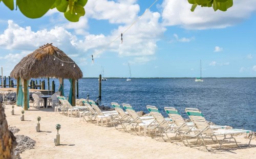 A sandy beach with lounge chairs, a thatched gazebo, and sailboats in the water under a blue sky with fluffy clouds.