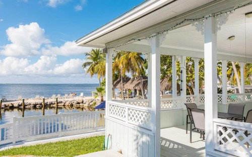A sunny waterfront view from a porch with palm trees and a dock in the background.
