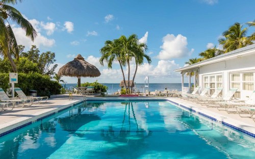 A serene pool area with lounge chairs, palm trees, and a thatched umbrella, overlooking a beach and ocean under a blue sky.