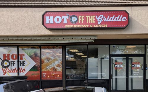 Sign for "Hot Off The Griddle" restaurant, featuring breakfast and lunch, with large windows and a welcoming entrance.