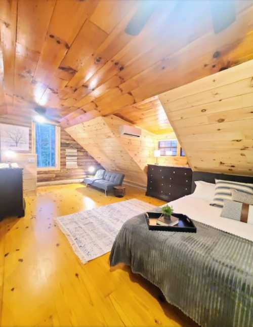 Cozy attic bedroom with wooden walls, a bed, a sofa, and a rug, featuring natural light from a window.