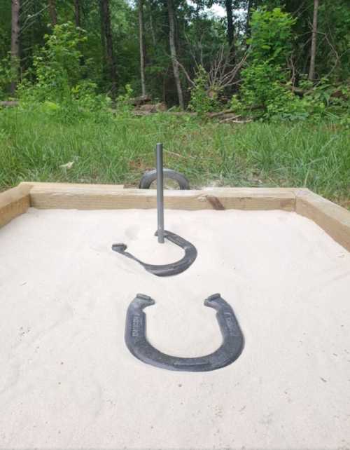 A horseshoe pit in a grassy area, featuring a wooden frame, sand, and two horseshoes near a vertical stake.