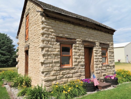 A small stone house with wooden windows and doors, surrounded by colorful flowers and American flags.