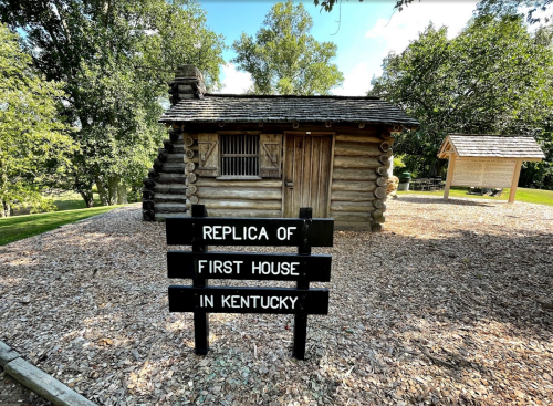 A log cabin with a sign reading "Replica of First House in Kentucky," surrounded by trees and gravel.