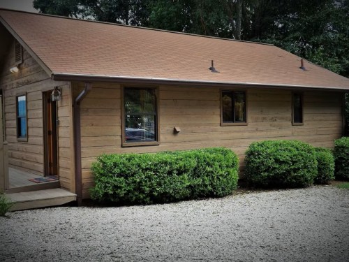 A wooden cabin with a brown roof, surrounded by green bushes and gravel, set in a wooded area.