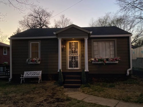 A cozy brown house with a white door, flower boxes, and a small bench in front, illuminated at dusk.