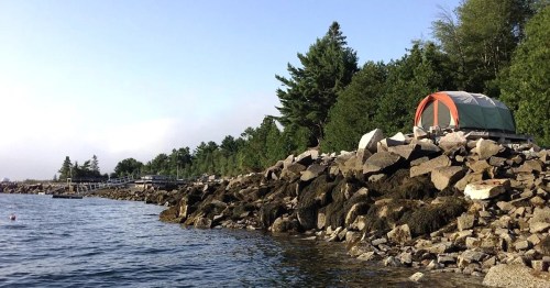 A tent on rocky shore near calm water, surrounded by trees and a distant pier under a clear sky.