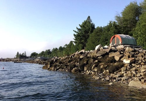 A tent set up on rocky shorelines beside calm water, surrounded by trees and a distant wooden pier.