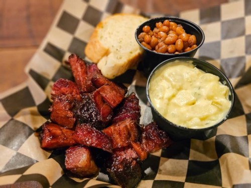 A plate of smoked meat chunks, baked beans, creamy potato salad, and garlic bread on a checkered paper.