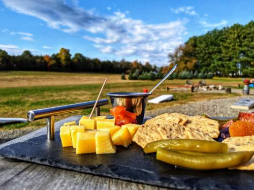 A cheese platter with assorted cheeses, pickles, and a small bowl, set against a scenic outdoor landscape.