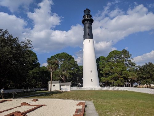 A tall black and white lighthouse surrounded by trees and a sandy area under a partly cloudy sky.