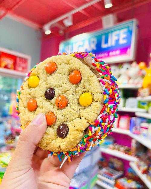 A hand holding a colorful cookie sandwich with sprinkles and candy pieces, set against a vibrant store background.