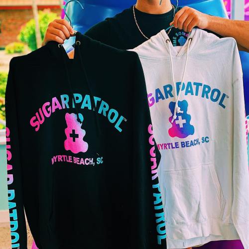 A person holds two hoodies, one black and one white, both featuring the "Sugar Patrol" logo in colorful text.