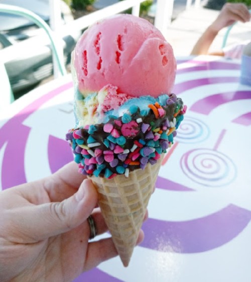 A hand holding a colorful ice cream cone with pink and yellow scoops topped with vibrant sprinkles.