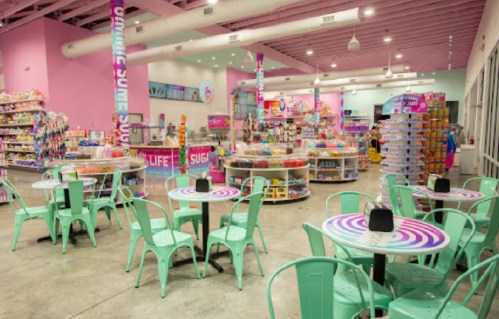 Brightly colored candy store interior with pastel decor, circular tables, and shelves filled with sweets and treats.