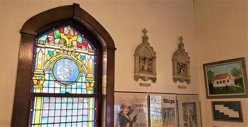 Stained glass window with colorful design, flanked by wall-mounted sculptures and framed artwork in a museum setting.