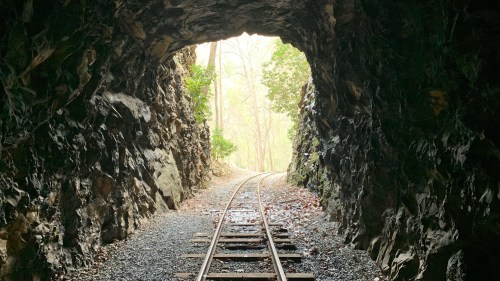 View from inside a rocky tunnel, with railroad tracks leading out to a bright, tree-lined path.