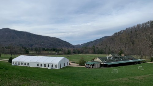 A large white tent and a building sit on a grassy field, surrounded by mountains under a cloudy sky.