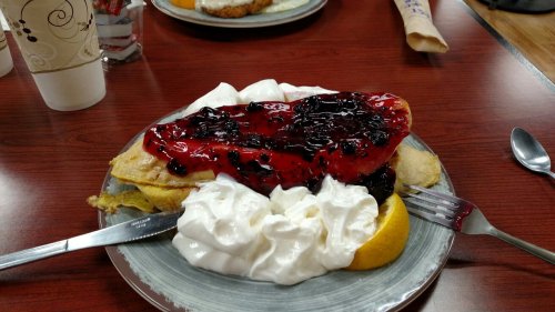 A plate of pancakes topped with berry jam, whipped cream, and a slice of lemon, served on a wooden table.