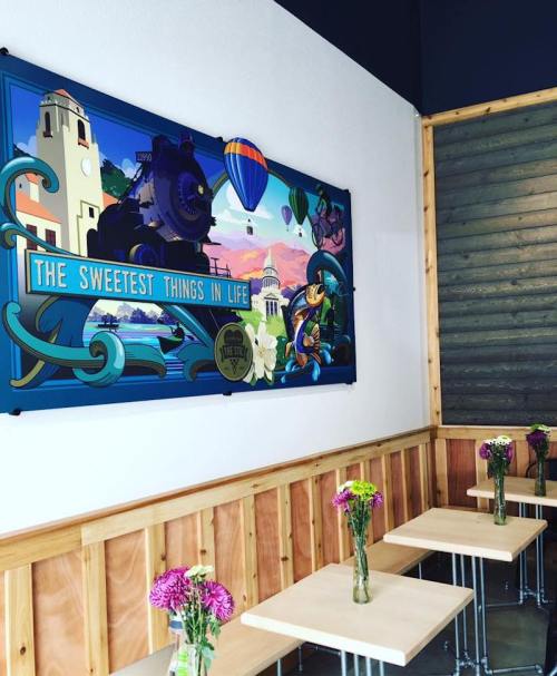Colorful mural titled "The Sweetest Things in Life" on a cafe wall, with wooden tables and flower centerpieces.