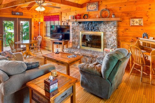 Cozy cabin living room with wooden walls, a stone fireplace, comfortable seating, and a view of the outdoors.