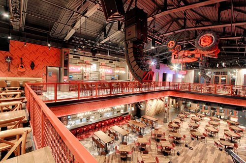 A spacious restaurant interior with wooden tables, a stage, and a vintage car hanging from the ceiling.