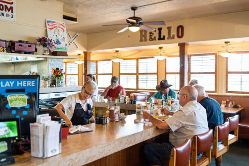 A diner scene with a waitress serving food to customers at the counter, bright windows, and a cozy atmosphere.