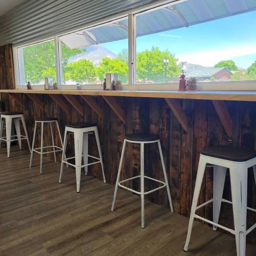 A row of white bar stools against a wooden counter, with large windows letting in natural light and greenery outside.