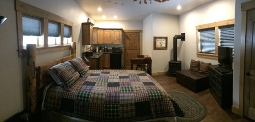 Cozy bedroom with a wooden bed, plaid bedding, kitchenette, and a small seating area. Warm, rustic decor.